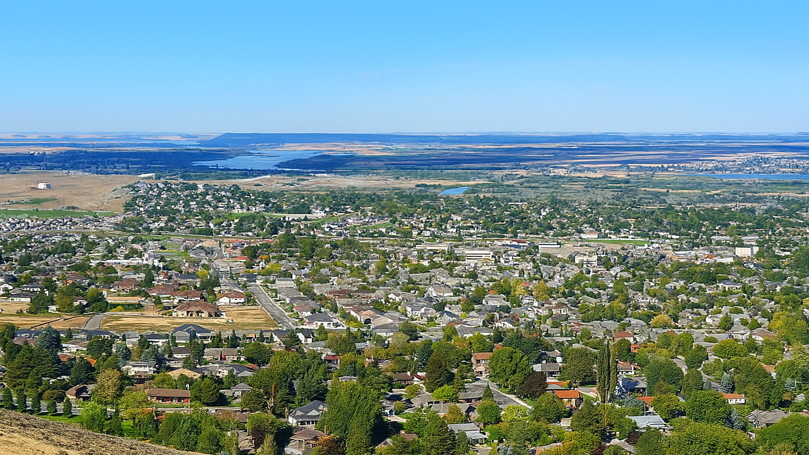 Panoramic view of Pasco, Washington, showing residential neighborhoods, tree-lined streets, and the Columbia River in the distance under a clear blue sky.
