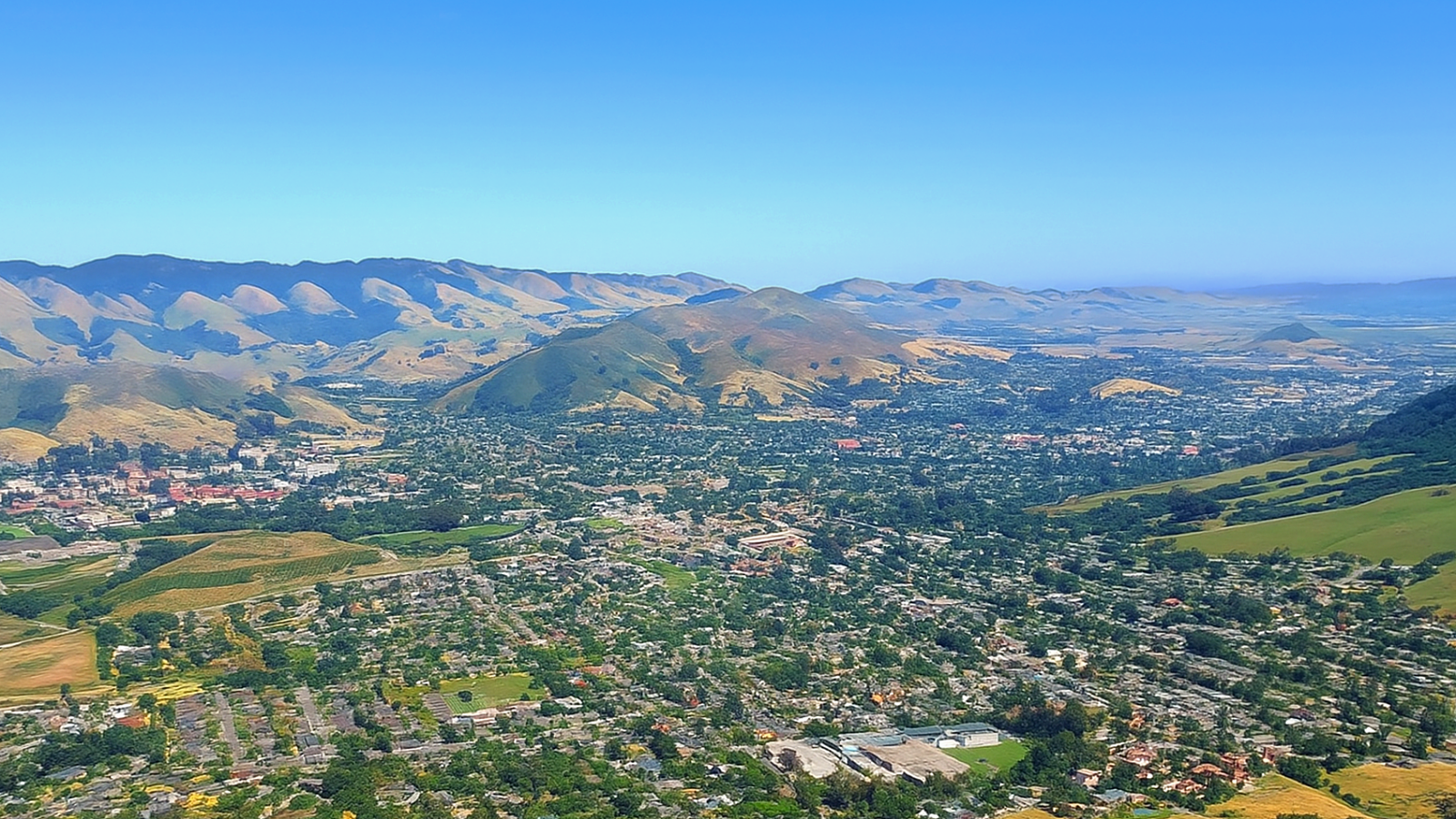 A panoramic view of San Luis Obispo, California, showing the city nestled among gentle green hills under a clear blue sky.